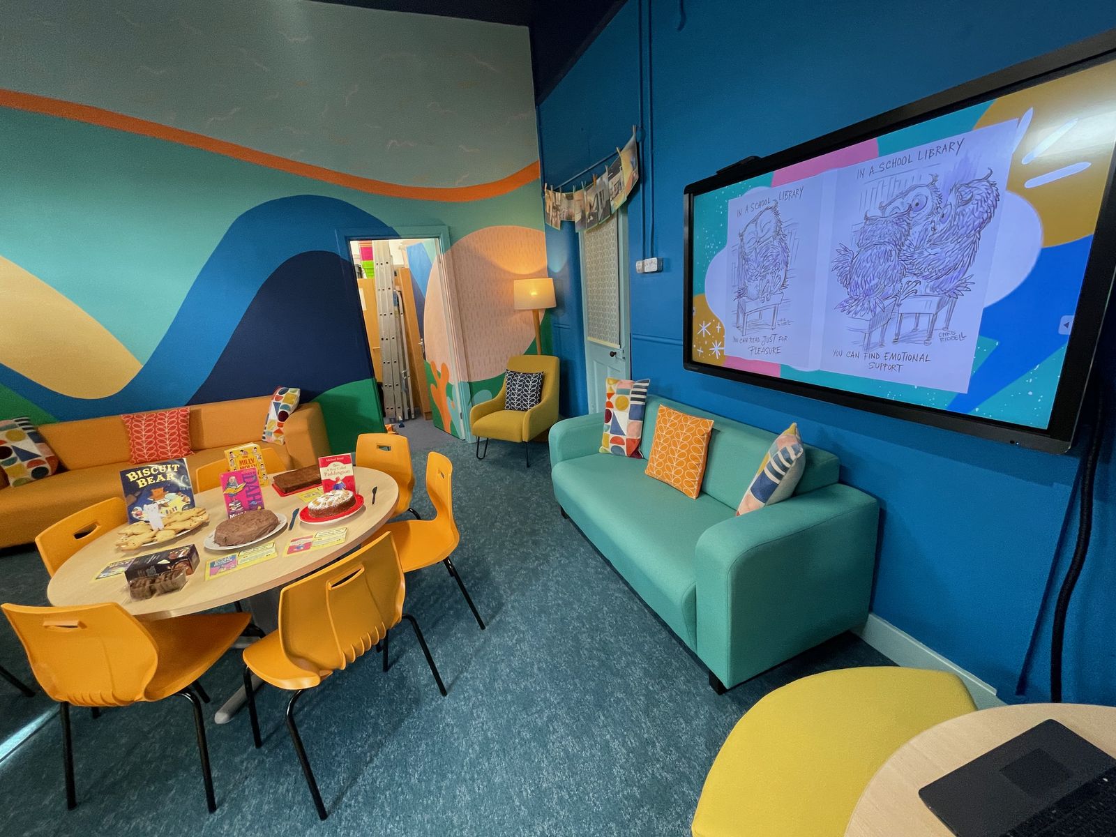 The Caughlin Library ready for its opening: a round table of cakes and books, orange chairs, turquoise sofas and a wave mural on the wall