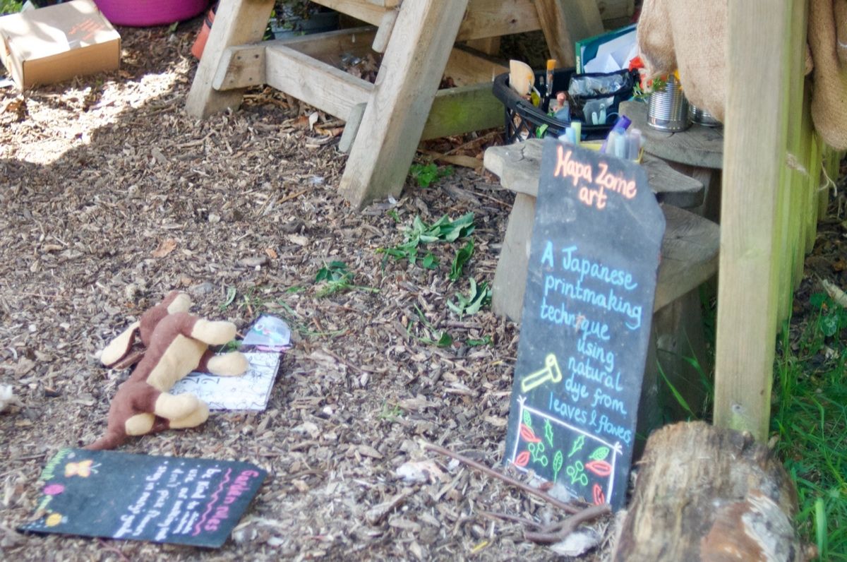 A chalkboard sign propped against a wooden trestle in Mary's Garden, reading: 'Hapa Zome art — A Japanese printmaking technique using natural dye from leaves and flowers'. Felt-cut animals and a small label-chalkboard sit on the mulch beside it.