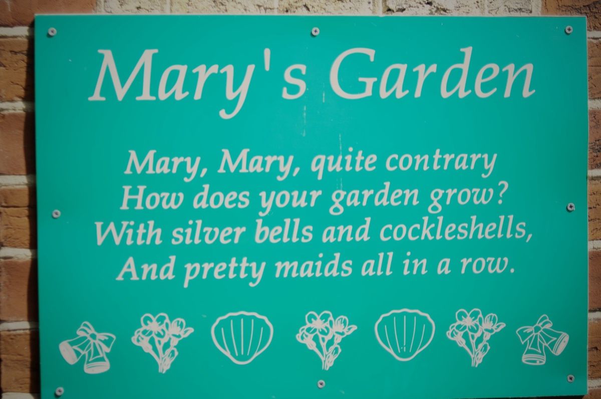 The Mary's Garden sign on the school wall — a brand-teal painted board with the title 'Mary's Garden', the full four-line nursery rhyme, and decorative line-drawings of silver bells, flowers and cockle shells along the bottom edge.