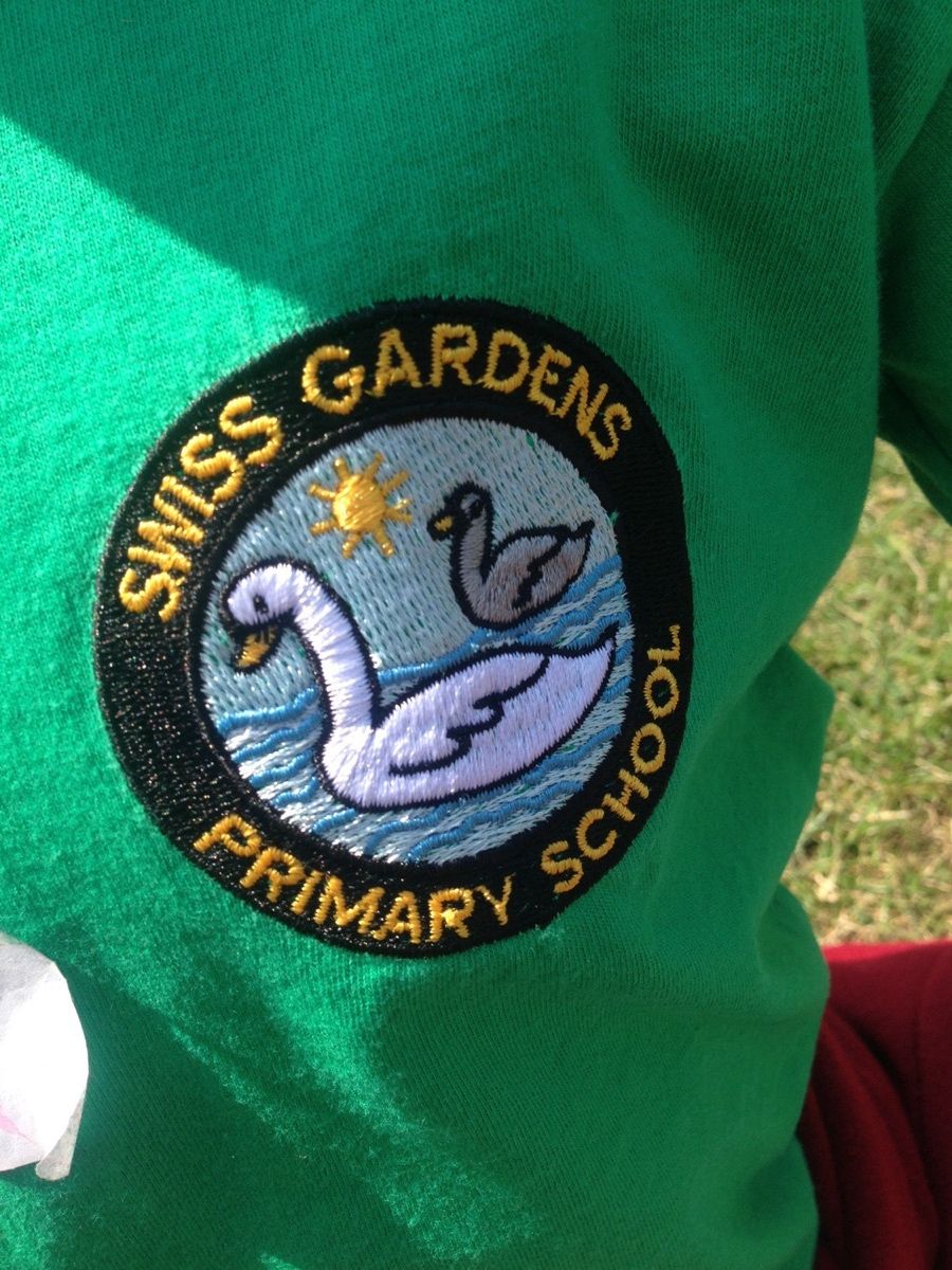 Close-up of the Swiss Gardens school crest embroidered on a green house t-shirt, showing two swans on water beneath a sun